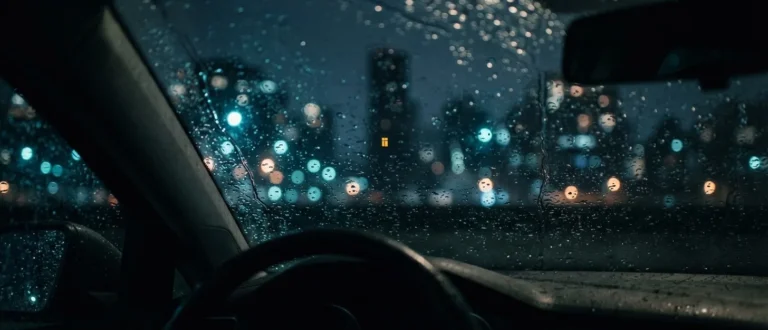 Vista de dentro de um carro à noite, com gotas de chuva no para-brisa e luzes da cidade desfocadas ao fundo